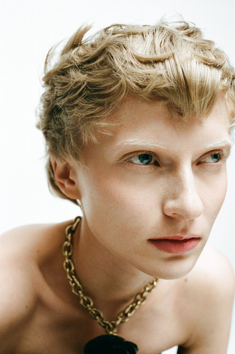 Close-up portrait of a young adult female with short blonde wavy hair, light complexion, wearing a gold chain necklace against a white background.