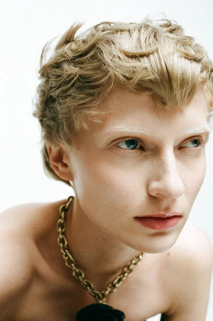 Close-up portrait of a young adult female with short blonde wavy hair, light complexion, wearing a gold chain necklace against a white background.
