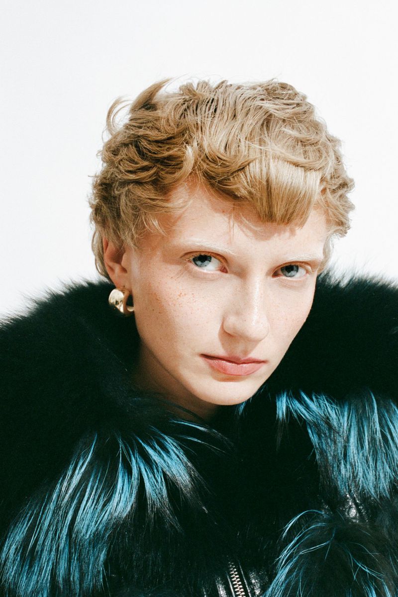 Close-up shot of a blond adult woman wearing a black and turquoise fur coat and gold earrings, looking directly at the camera in a studio.