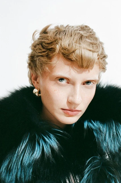 Close-up shot of a blond adult woman wearing a black and turquoise fur coat and gold earrings, looking directly at the camera in a studio.