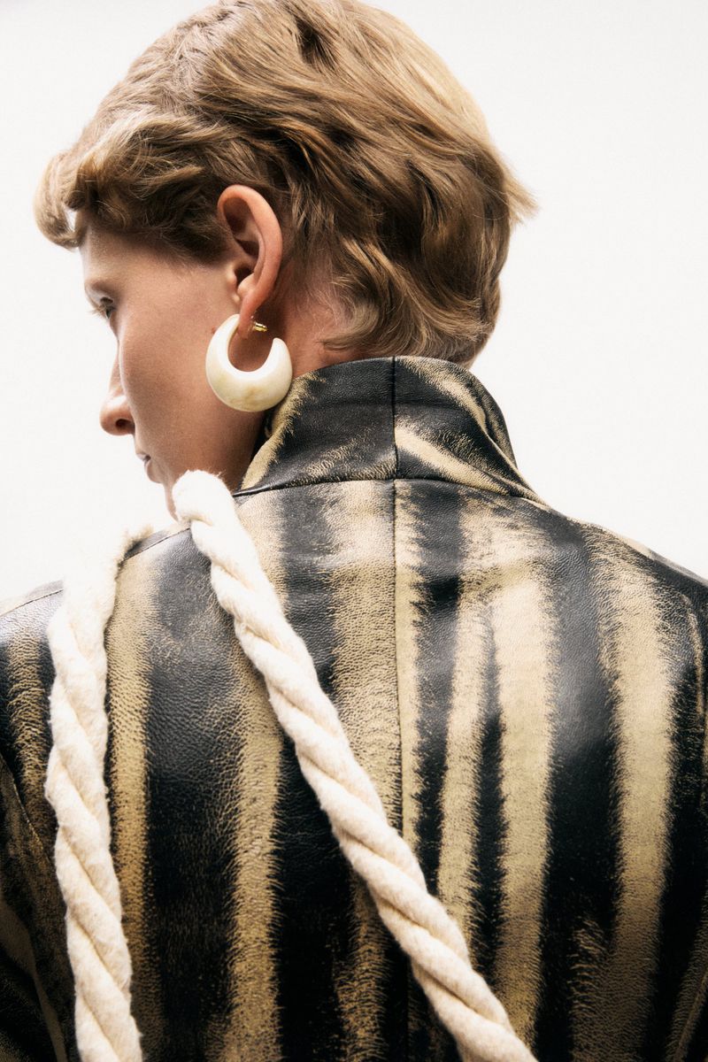 Back view of an adult female with short hair wearing a black and tan striped jacket with a white rope strap over her shoulder.