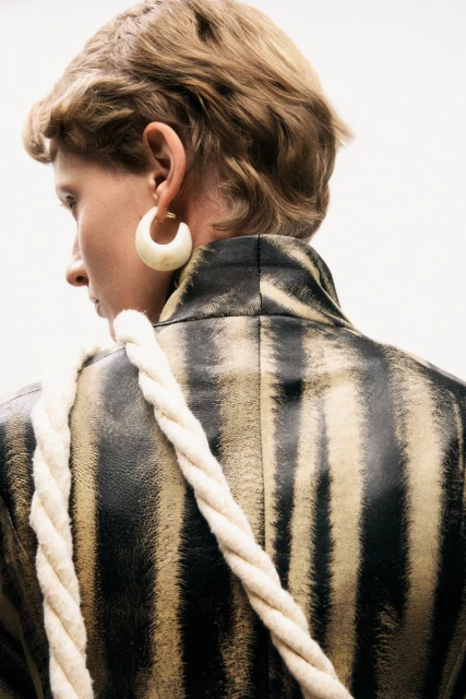 Back view of an adult female with short hair wearing a black and tan striped jacket with a white rope strap over her shoulder.