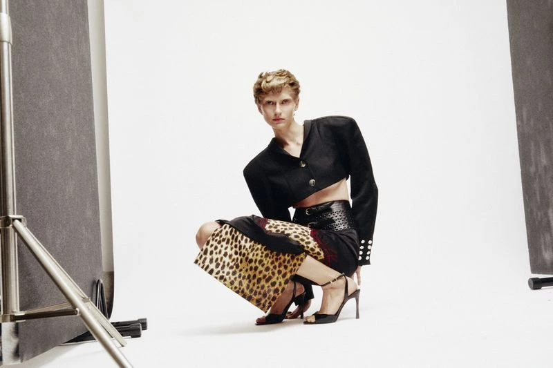 Adult woman with short hair poses in a black crop top and printed skirt, crouching in a photo studio with a light backdrop.