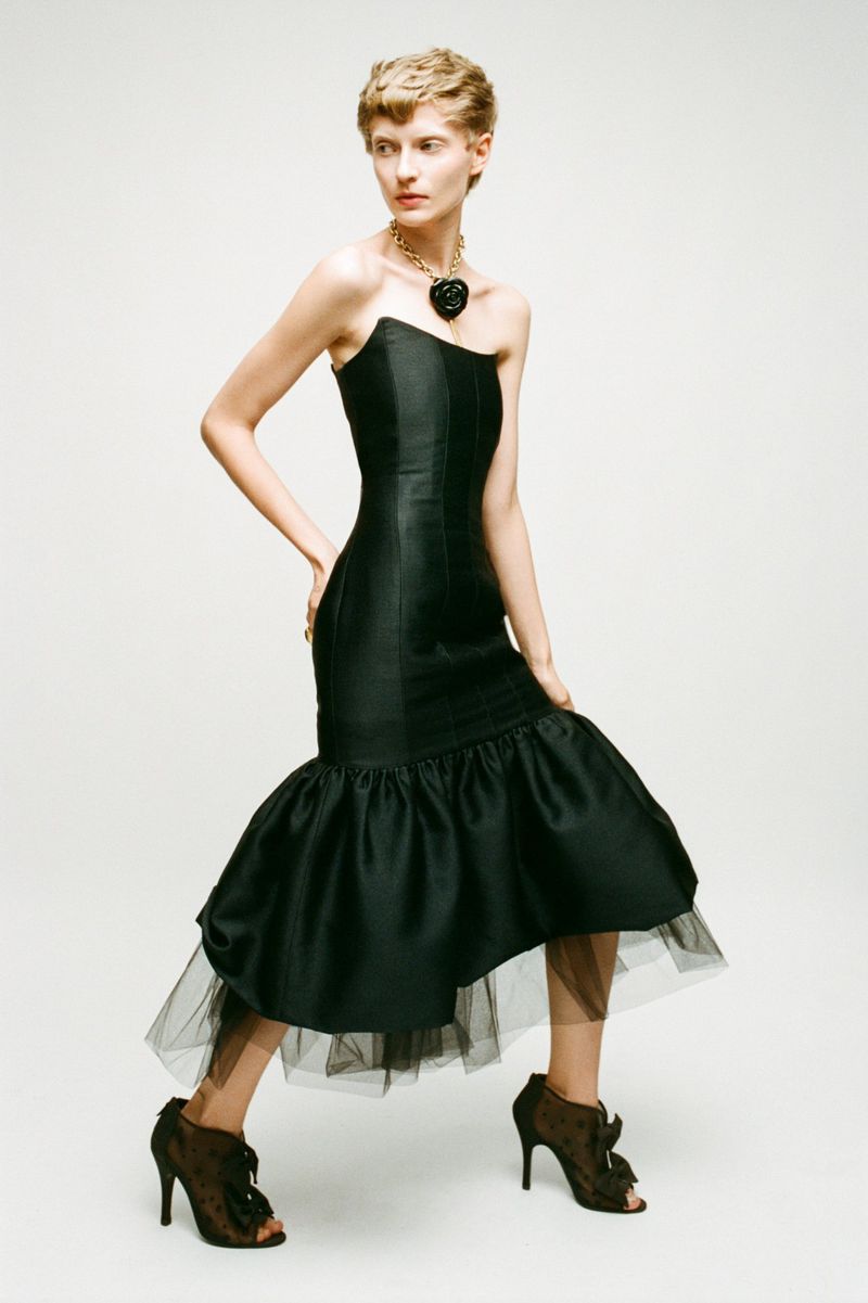 A slender woman with short blonde hair poses in a black strapless gown with ruffled hem, accessorized with a necklace, against a neutral backdrop.