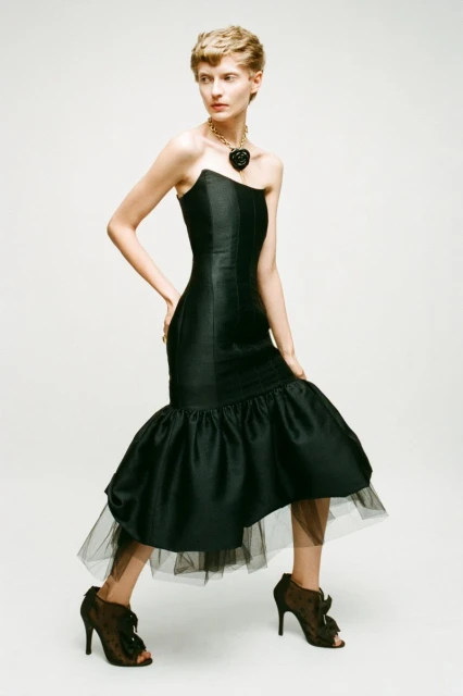 A slender woman with short blonde hair poses in a black strapless gown with ruffled hem, accessorized with a necklace, against a neutral backdrop.