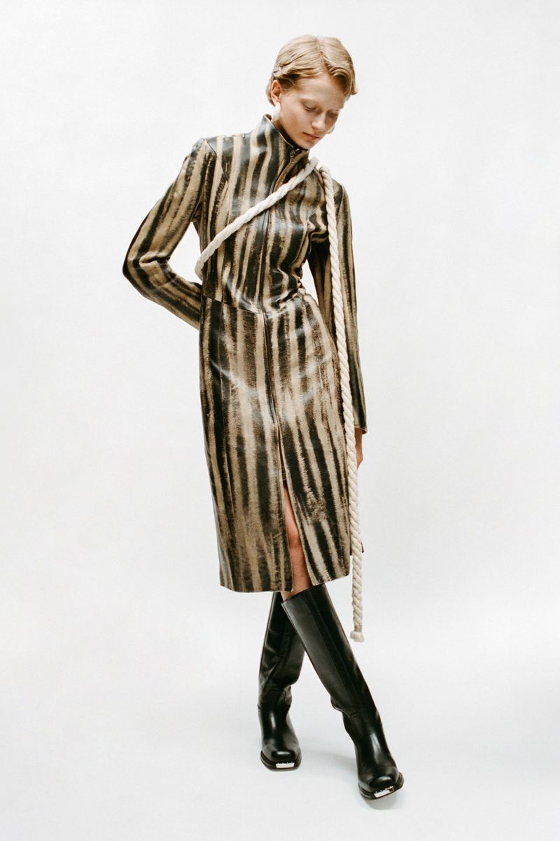 Adult female fashion model wearing a black and brown striped dress with black boots, posing in front of a white backdrop in a studio.