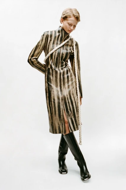 Adult female fashion model wearing a black and brown striped dress with black boots, posing in front of a white backdrop in a studio.