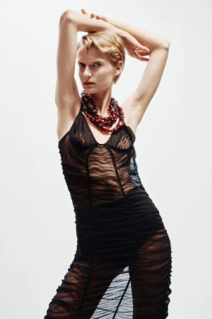 Fashion portrait of a blonde woman wearing a black dress and statement necklace in a studio with a neutral, light background.