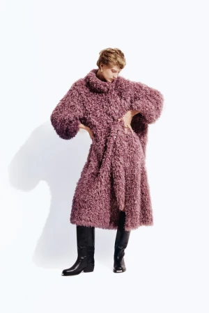 Model wearing a fuzzy mauve textured coat and black boots standing against a white backdrop