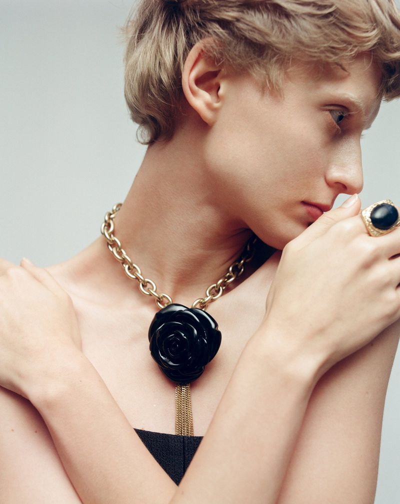 Woman wears a gold chain necklace featuring a large black rose pendant and a large black gemstone ring in a studio setting.