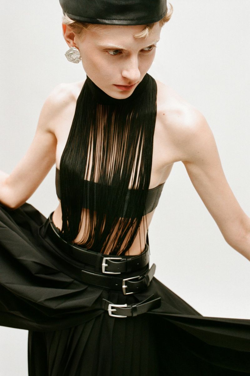 Fashion model in black dress with fringe, leather cap, and round earrings in a studio setting, showcasing editorial style and design.