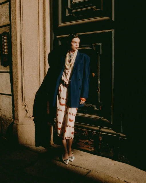 A stylish woman in a blue blazer and printed dress poses in front of a dark door with architectural details, bathed in warm light.