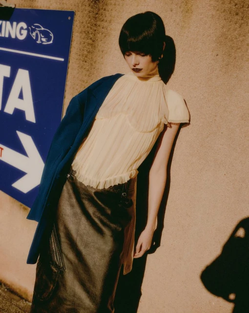 A fair-skinned woman with dark hair poses in a cream-colored top and brown skirt, standing in front of a weathered blue sign.