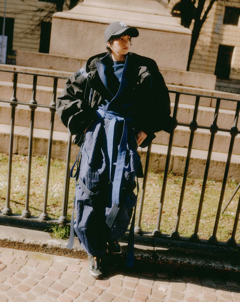 A woman in an oversized black coat and blue layered skirt poses in front of a wrought-iron fence on a brick sidewalk during daytime.