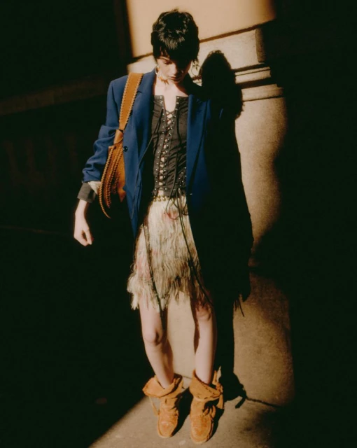 A woman in a blue blazer, corset top, and brown boots stands in a shadowy setting with a brown purse; fashion editorial shot.
