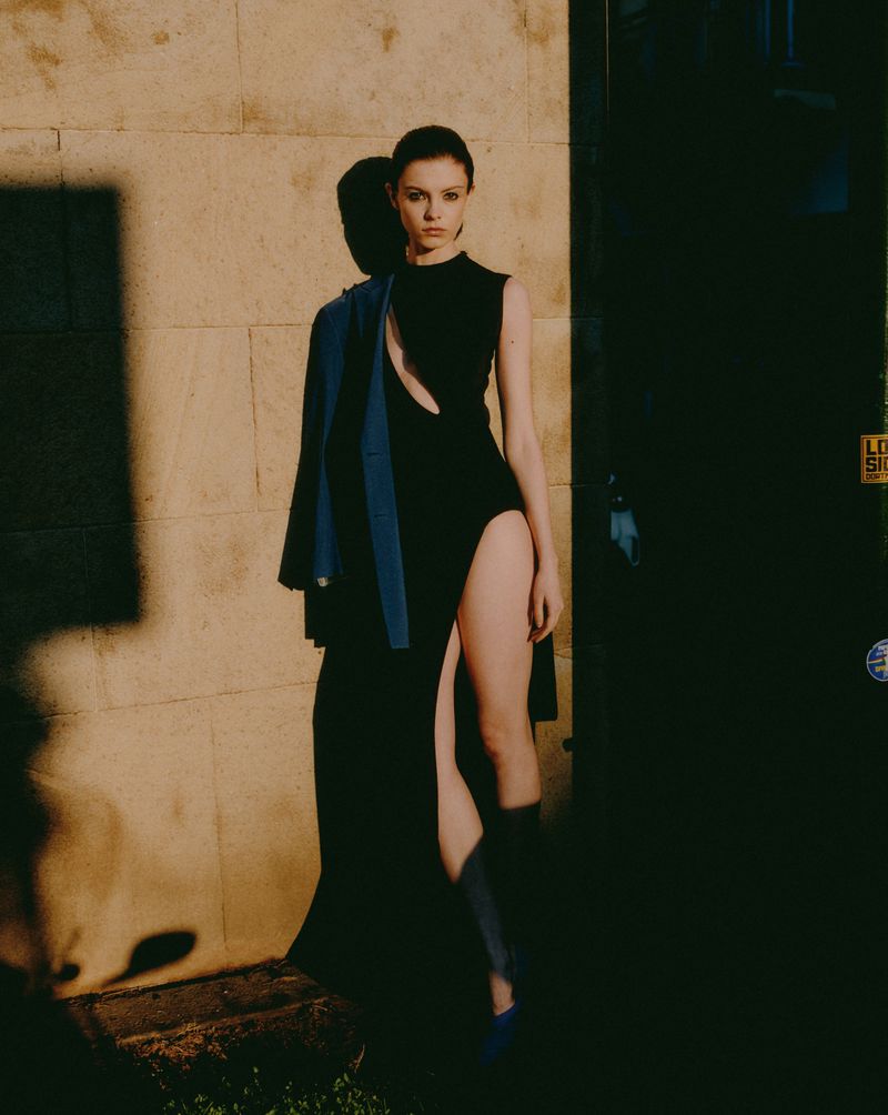 A fashionable woman in a black dress stands outdoors in dappled sunlight with a blue jacket slung over her shoulder.