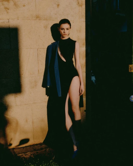 A fashionable woman in a black dress stands outdoors in dappled sunlight with a blue jacket slung over her shoulder.