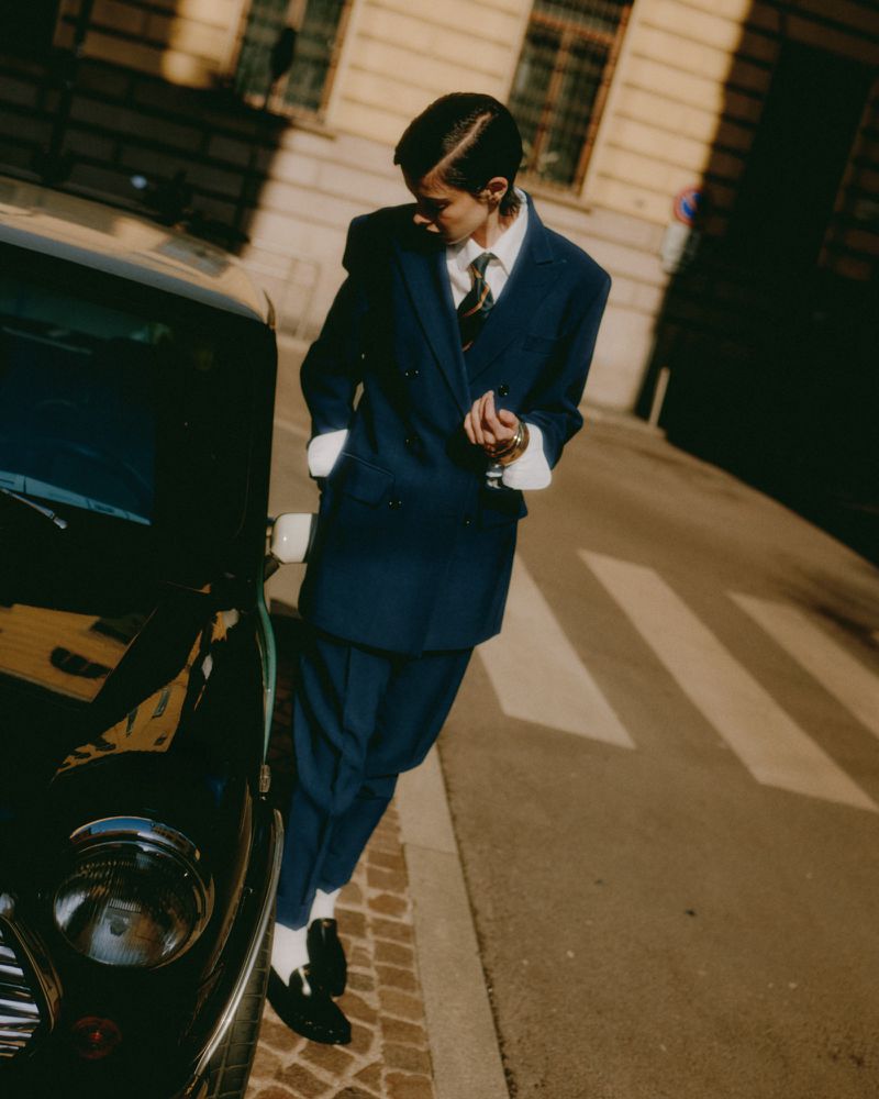 Stylish young adult poses in a navy suit alongside a vintage car on a city street, bathed in warm sunlight. Classic menswear fashion.