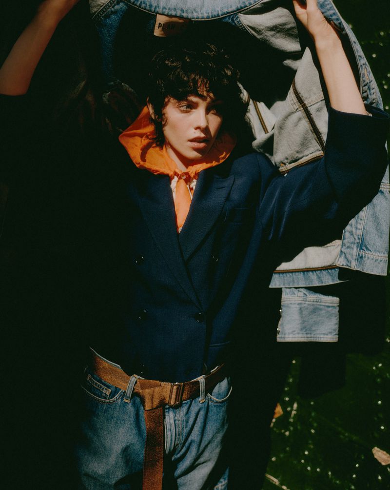 Brunette woman in blazer, jeans, and belt poses with denim jackets indoors with dark lighting and a gritty texture in a fashion shot.