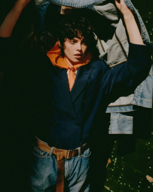 Brunette woman in blazer, jeans, and belt poses with denim jackets indoors with dark lighting and a gritty texture in a fashion shot.