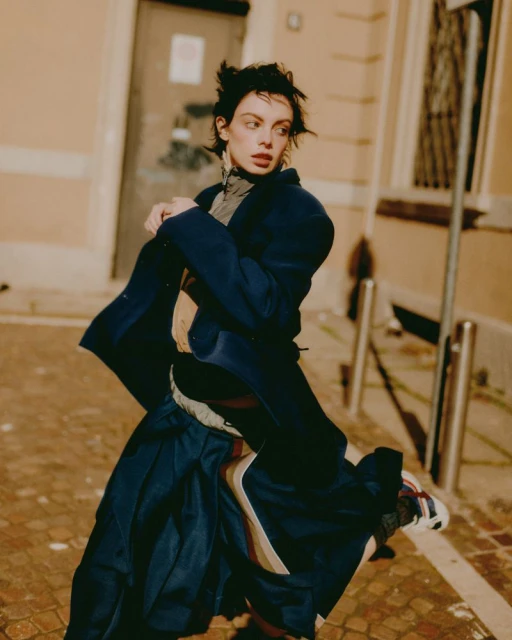 Woman in blue coat poses dynamically on urban street. She wears a stylish outfit, sneakers, and has short hair. Street photography aesthetic.