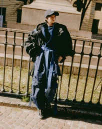 Model in layered blue garments wearing a black baseball cap standing outdoors by a metal fence and stone steps.