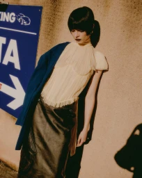 Model with dark hair wearing a pale yellow pleated top and dark skirt leaning against a concrete wall outdoors