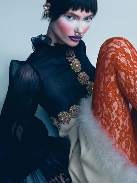 Close-up of a woman with creative makeup, styled dark hair, and orange lace leggings against a light background in a studio setting.