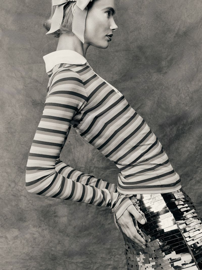 Woman in striped shirt and metallic skirt posing against a textured background in a studio shot. Fashion editorial, portrait orientation.