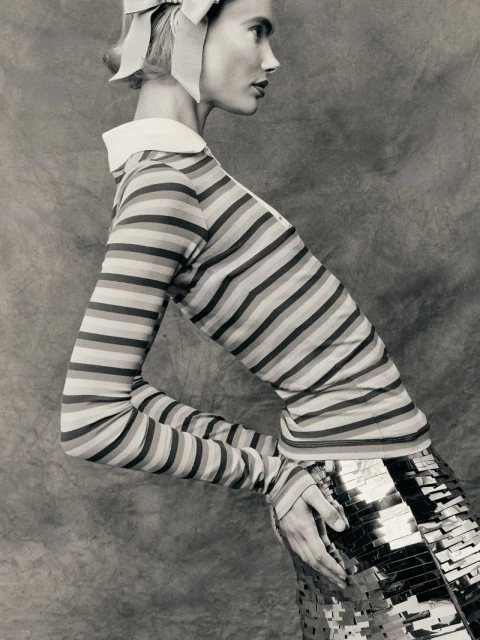 Woman in striped shirt and metallic skirt posing against a textured background in a studio shot. Fashion editorial, portrait orientation.