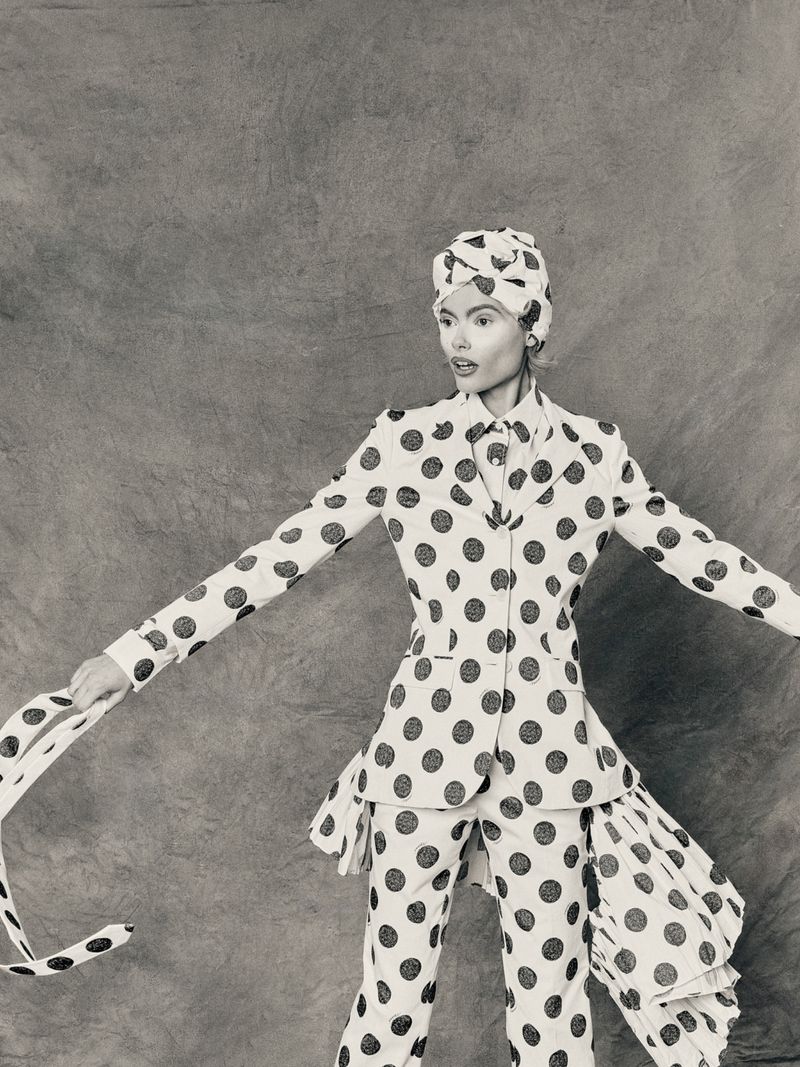 Monochrome fashion shot of a woman in a polka-dot suit and head wrap with arms outstretched against a textured gray backdrop.