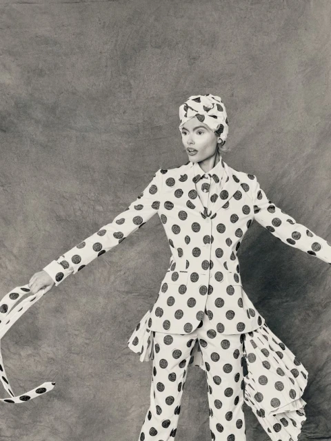 Monochrome fashion shot of a woman in a polka-dot suit and head wrap with arms outstretched against a textured gray backdrop.
