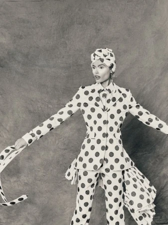 Monochrome fashion shot of a woman in a polka-dot suit and head wrap with arms outstretched against a textured gray backdrop.