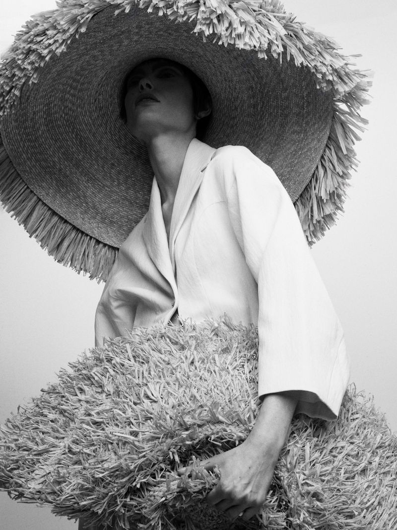 A woman wearing a straw hat and jacket, holding a straw bag, looks up in this black and white, medium shot studio fashion portrait.