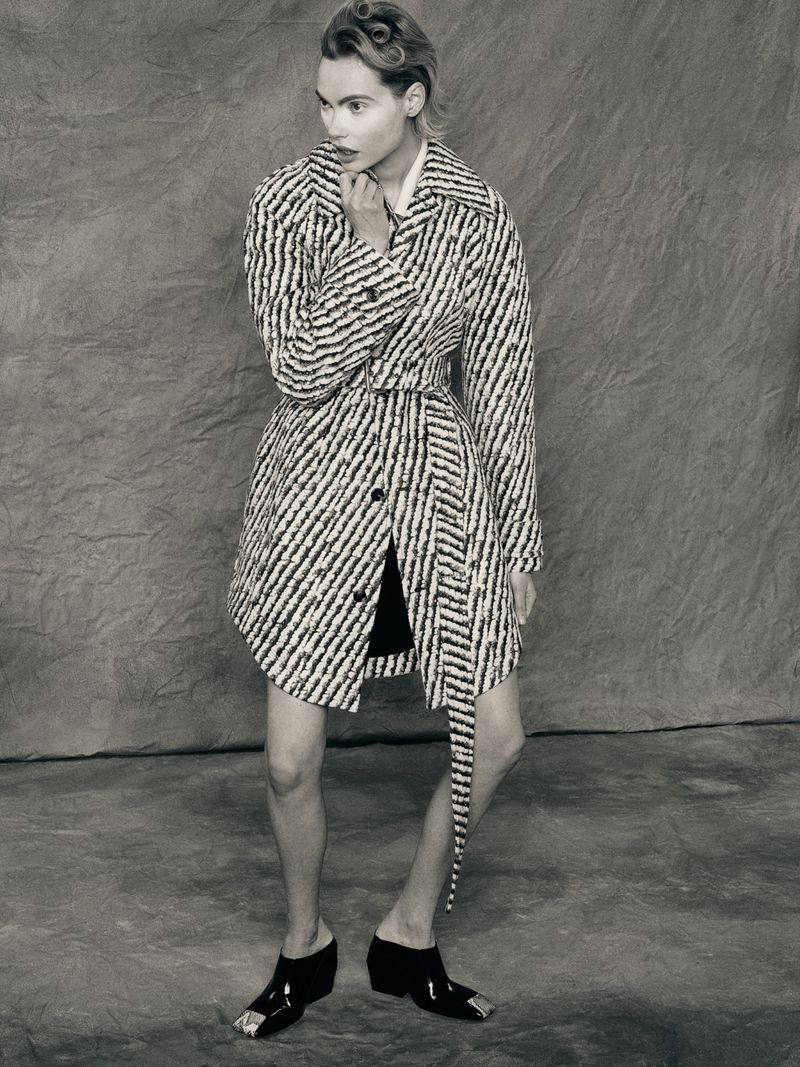 A woman wearing a striped coat and metallic-toe shoes poses against a textured backdrop in a studio fashion shot.