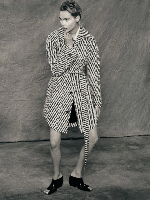 A woman wearing a striped coat and metallic-toe shoes poses against a textured backdrop in a studio fashion shot.