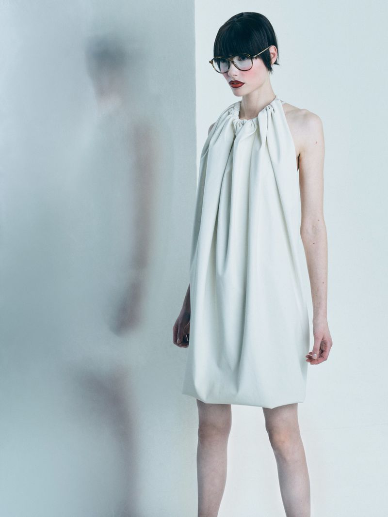Studio shot of a young adult female fashion model wearing a white dress and glasses, posing against a white background next to a frosted glass panel.