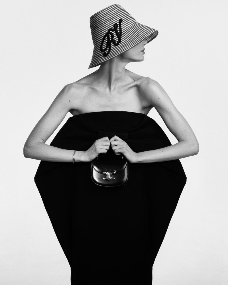 Monochrome fashion shot of a woman in a striped hat holding a black purse, posing against a white background in a studio.