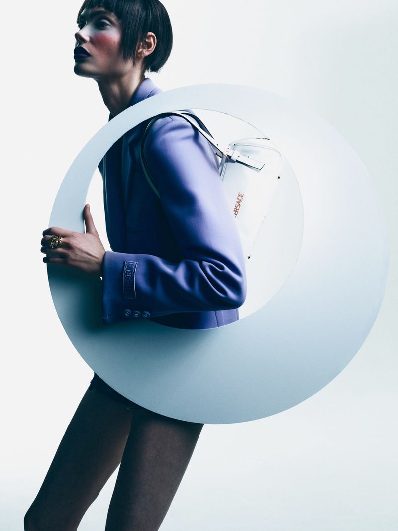 Fashion shot of a person in a purple jacket, dark lipstick and short hair, posing with a large circular object. 