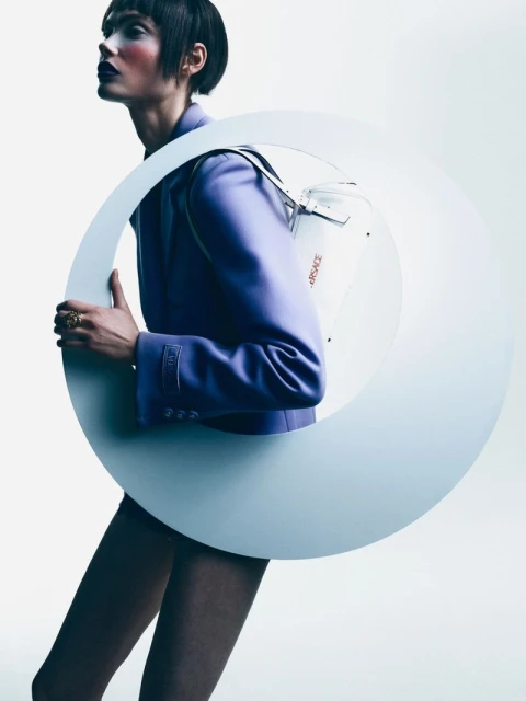 Fashion shot of a person in a purple jacket, dark lipstick and short hair, posing with a large circular object. 