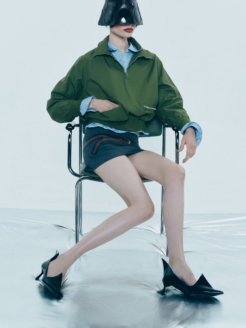 A fashion image featuring an adult female model wearing a stylish green jacket, gray skirt, and black heels, seated on a modern chair.