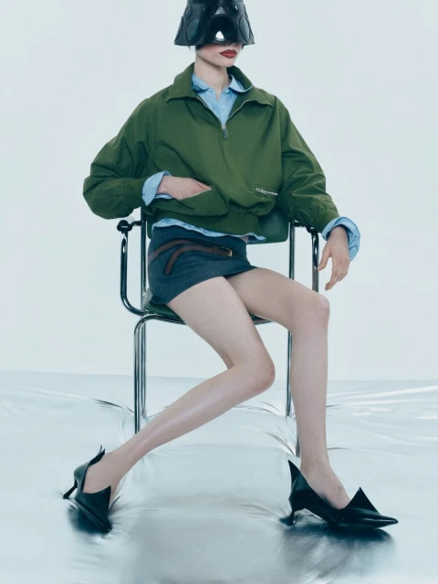 A fashion image featuring an adult female model wearing a stylish green jacket, gray skirt, and black heels, seated on a modern chair.