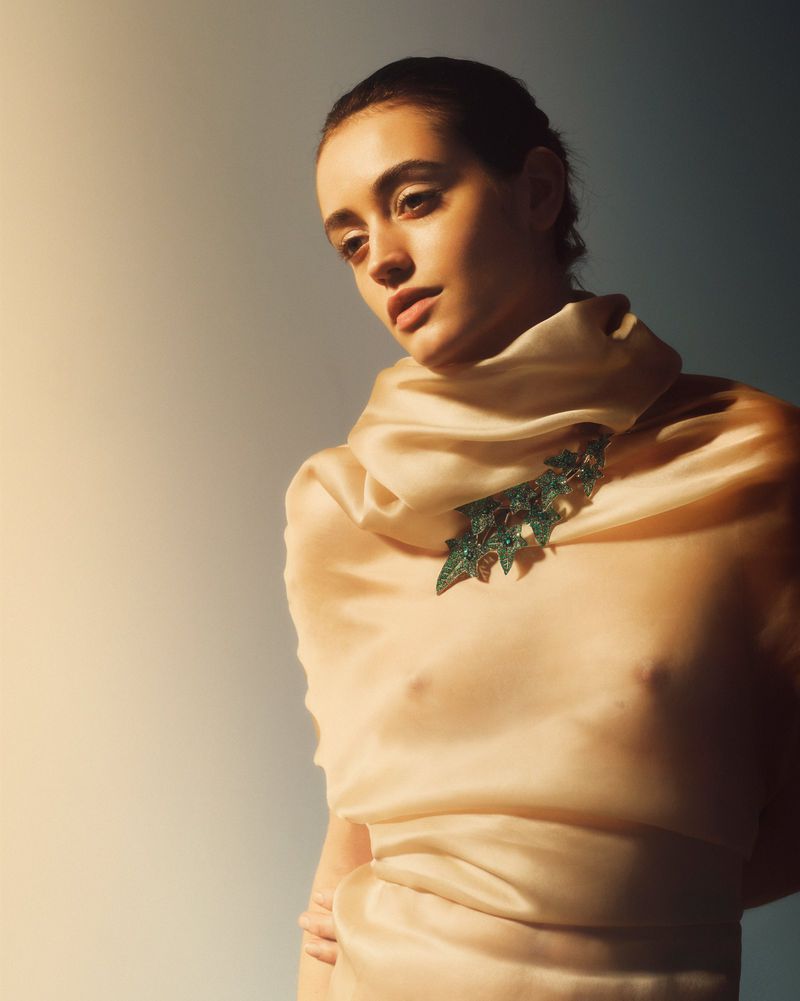 Adult woman with fair skin poses in a cream-colored draped garment with green leaf-shaped jeweled brooch. Low angle. Studio lighting.