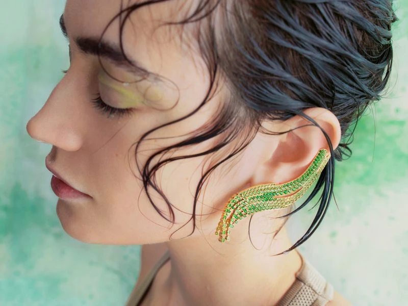 Profile of a woman with green and gold ear cuff jewelry. Wet hair is styled back, showcasing jewelry on her ear against a mottled green backdrop.