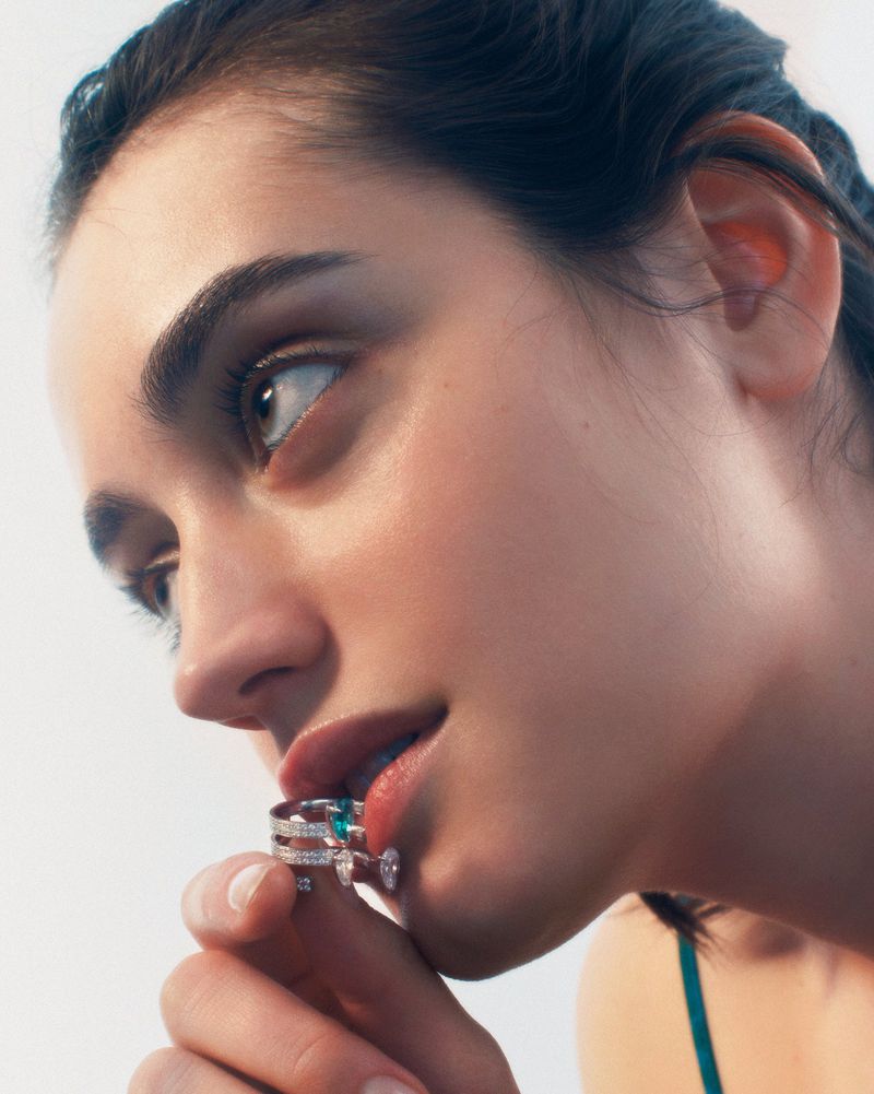 Close-up of a fair-skinned woman with dark hair and neutral expression holding jeweled rings to her lips in a brightly lit studio setting.