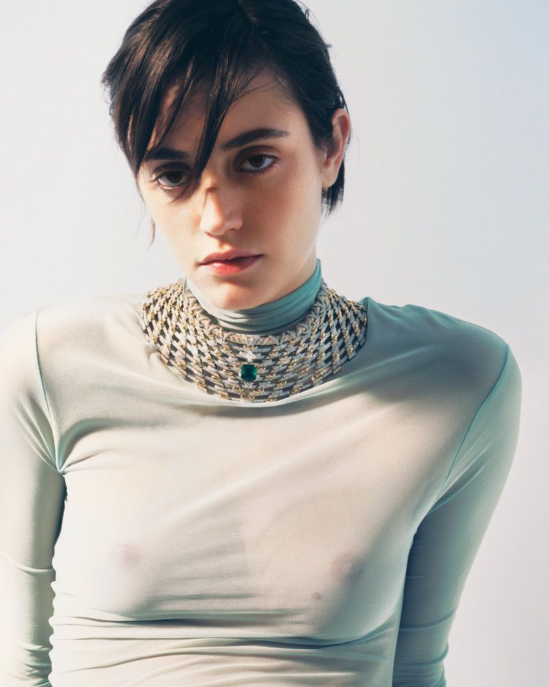 Elegant portrait of a woman with short dark hair wearing a sheer top and a statement collar necklace with an emerald green stone.