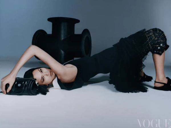 Fashion editorial shot of an adult female model in a black dress, posing on a white surface with a black bag and a sculptural pipe.