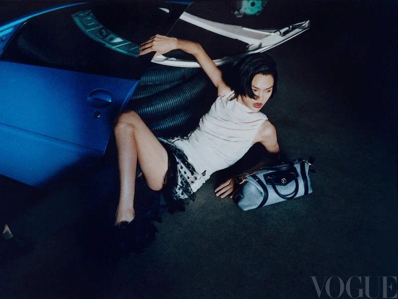 Vogue fashion editorial featuring an adult female model in a light-colored dress, lounging next to a blue sportscar and a handbag in a studio.