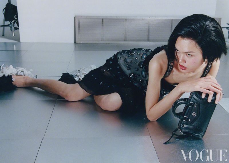 A woman in a black dress reclines on a silver floor with a black purse, Vogue logo visible. Fashion editorial, studio lighting.