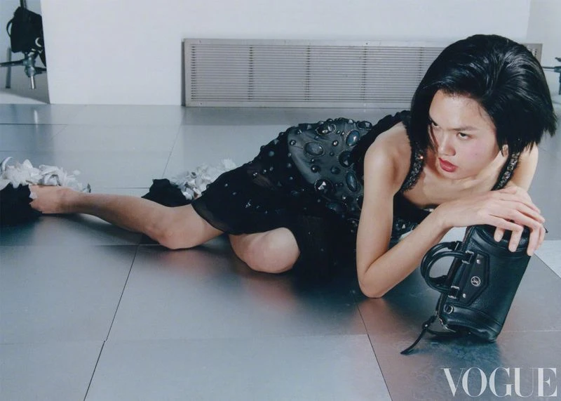 A woman in a black dress reclines on a silver floor with a black purse, Vogue logo visible. Fashion editorial, studio lighting.